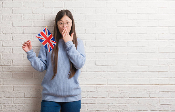 Young Chinese woman over brick wall holding flag of England cover mouth with hand shocked with shame for mistake, expression of fear, scared in silence, secret concept - Photo, Image