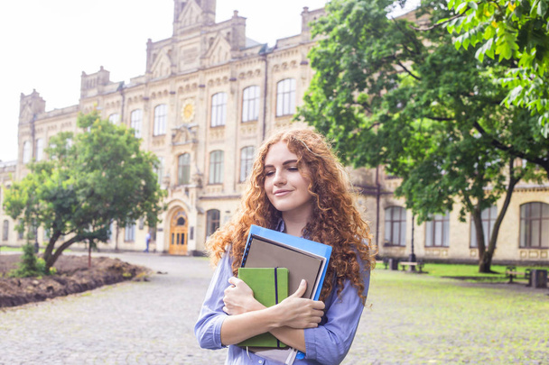 Una chica de pelo rojo rizado está de pie cerca del edificio de la universidad con materiales educativos, carpetas y un cuaderno para conferencias en sus manos. Concepto de estudio. Regreso a la escuela
 - Foto, Imagen