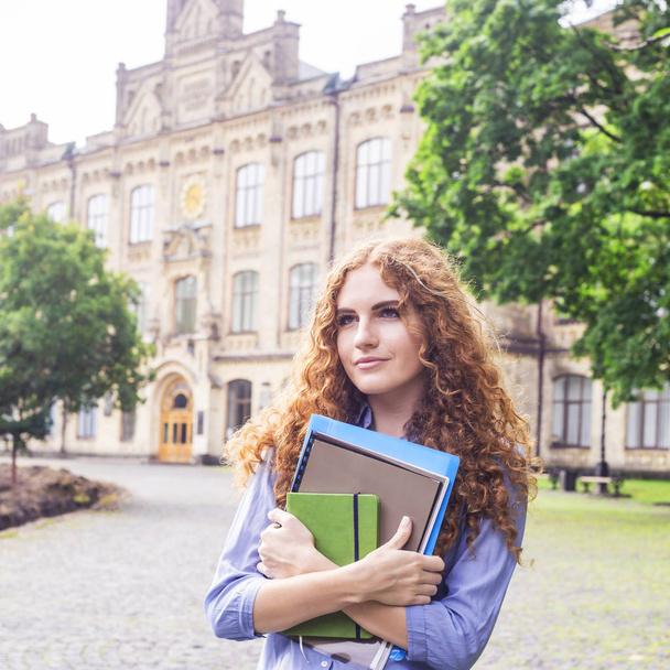 Uma menina ruiva encaracolada está de pé perto do prédio da universidade com materiais educacionais, pastas e um caderno para palestras em suas mãos. Conceito de estudo. De volta à escola
 - Foto, Imagem