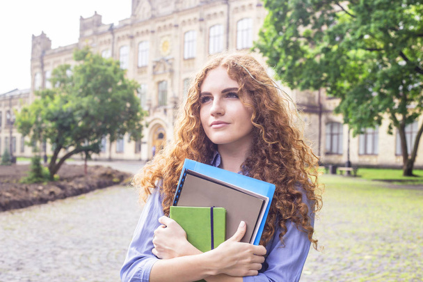Una chica de pelo rojo rizado está de pie cerca del edificio de la universidad con materiales educativos, carpetas y un cuaderno para conferencias en sus manos. Concepto de estudio. Regreso a la escuela
 - Foto, imagen