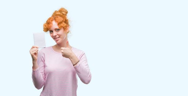 Young redhead woman holding blank notebook happy with big smile doing ok sign, thumb up with fingers, excellent sign - Foto, Imagen