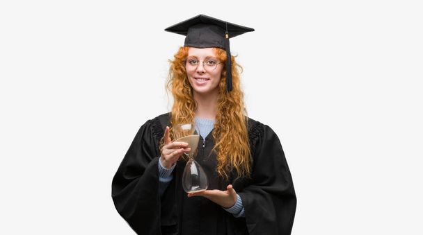 Молодая рыжая студентка в форме выпускника держит песочные часы со счастливым лицом стоя и улыбаясь с уверенной улыбкой показывая зубы
 - Фото, изображение