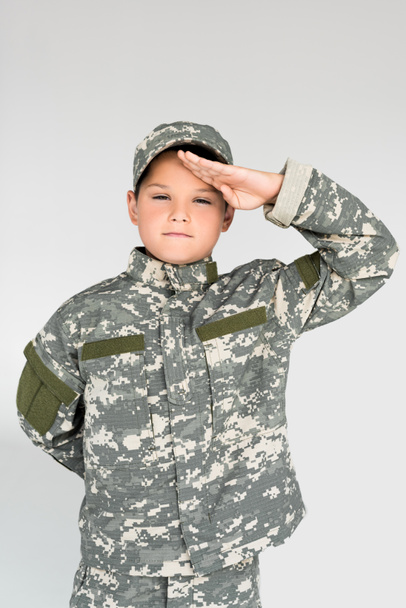 portrait of little kid in military uniform saluting on grey background - Фото, изображение