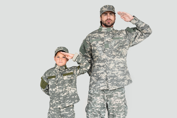 portrait of confident soldier and son in military uniforms saluting isolated on grey - Fotoğraf, Görsel