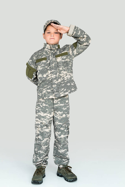 niño pequeño en uniforme militar mirando a la cámara y saludando sobre fondo gris
 - Foto, Imagen