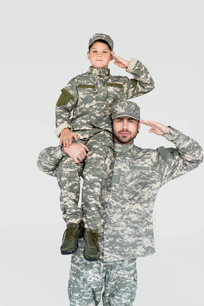 soldado sosteniendo hijo en uniforme militar en hombro y saludo aislado en gris
 - Foto, imagen