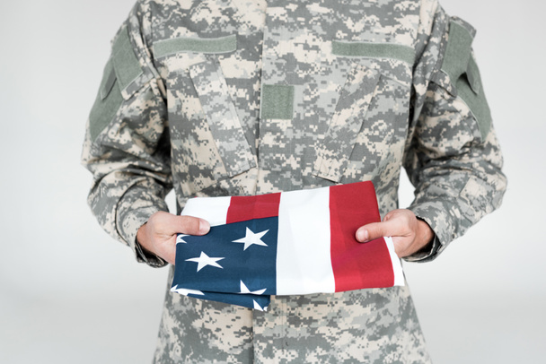 vista parcial del soldado en uniforme militar con bandera americana en las manos sobre fondo gris
 - Foto, Imagen
