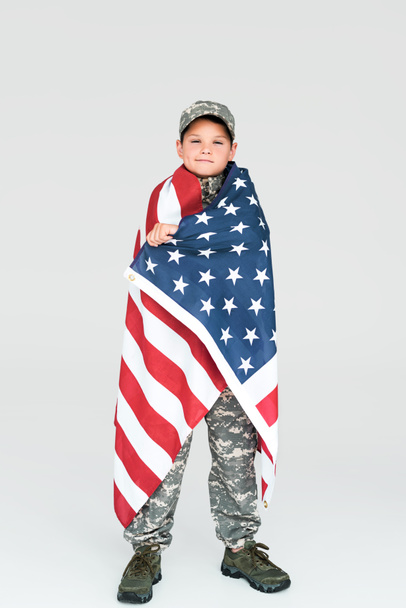 niño en uniforme militar cubierto con bandera americana mirando a la cámara sobre fondo gris
 - Foto, Imagen
