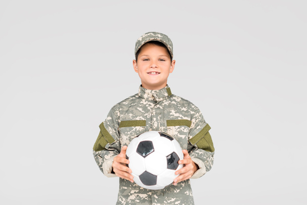 portret uśmiechający się dziecko w wojskowym mundurze z piłki nożnej w ręce na szarym tle - Zdjęcie, obraz