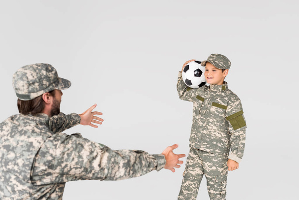 soldier with outstretched arms and smiling kid in camouflage clothing with soccer ball isolated on grey - Photo, Image