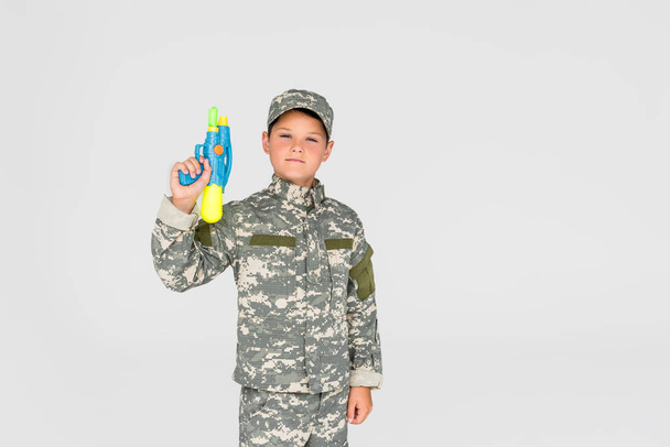 portrait of boy in camouflage clothing with toy water gun isolated on grey - Fotó, kép
