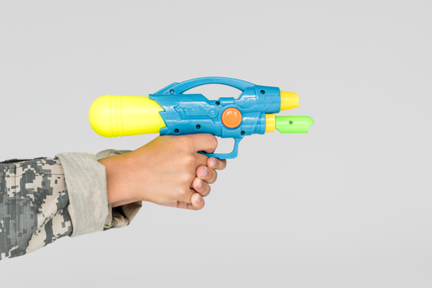 vista parcial de niño en ropa de camuflaje con pistola de agua de juguete en manos aisladas en gris
 - Foto, Imagen
