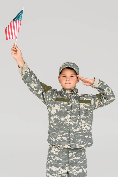portrait of boy in camouflage clothing saluting while holding american flagpole in hand isolated on grey - Foto, Imagen