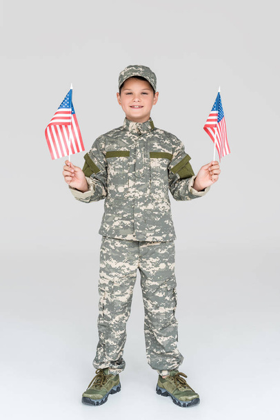 usmívající se dítě ve vojenské uniformě s americkým vlajkové stožáry v rukou při pohledu na fotoaparát na šedém pozadí - Fotografie, Obrázek
