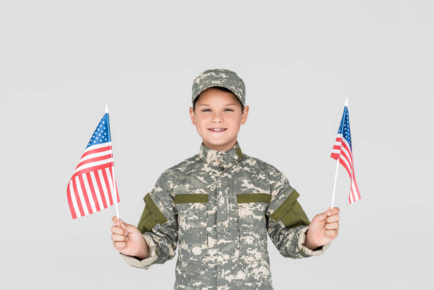 portrait of smiling child in military uniform with american flagpoles in hands isolated on grey - Fotoğraf, Görsel