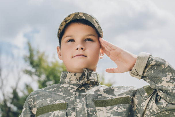 portrait of little boy in camouflage clothing saluting with cloudy sky on backdrop - Foto, Imagen