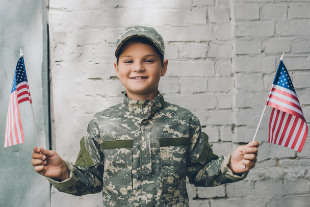 Porträt eines lächelnden Kindes in Tarnkleidung, das amerikanische Fahnenmasten mit grauer Ziegelwand vor dem Hintergrund hält - Foto, Bild