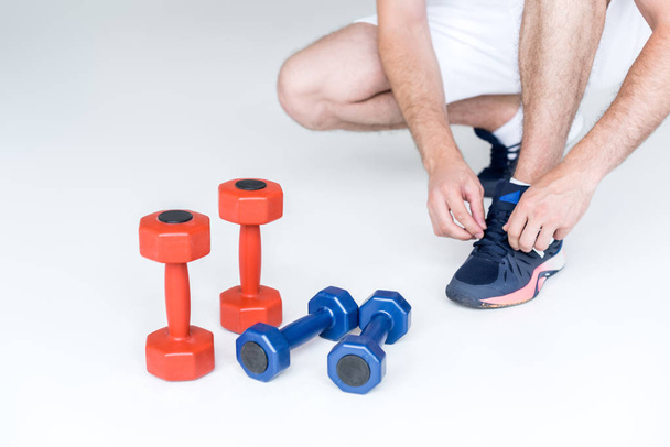 partial view of man tying shoelaces near red and blue dumbbells on grey backdrop - Photo, Image