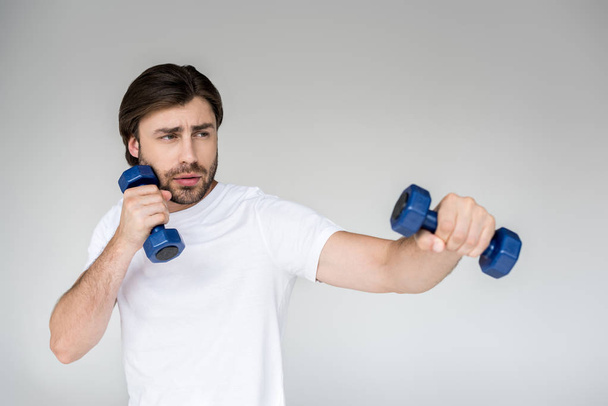 portrait of man in white shirt with blue dumbbells in hands exercising on grey backdrop - Фото, зображення