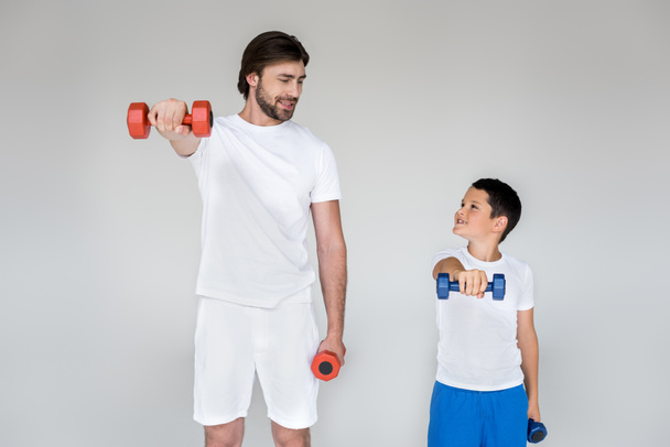 lachende vader en zoon in witte shirts kijken elkaar tijdens het trainen met halters op grijze achtergrond - Foto, afbeelding