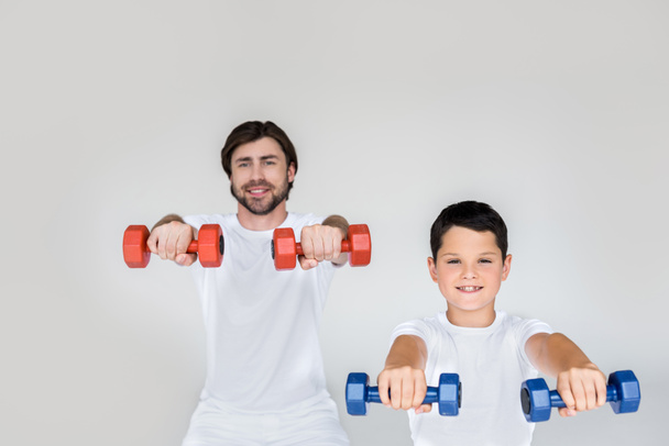 portrait of smiling father and son in white shirts exercising with dumbbells on grey background - Valokuva, kuva