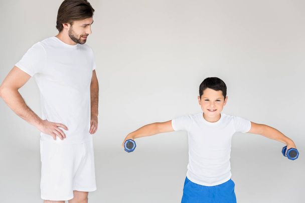 man standing akimbo and looking at son with dumbbells exercising on grey backdrop - Foto, immagini