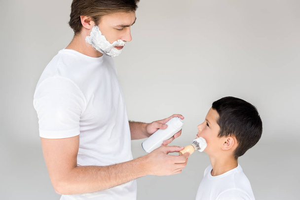 Вид сбоку отца с кисточкой, наносящей пену для бритья на лицо сыновей на сером фоне
 - Фото, изображение
