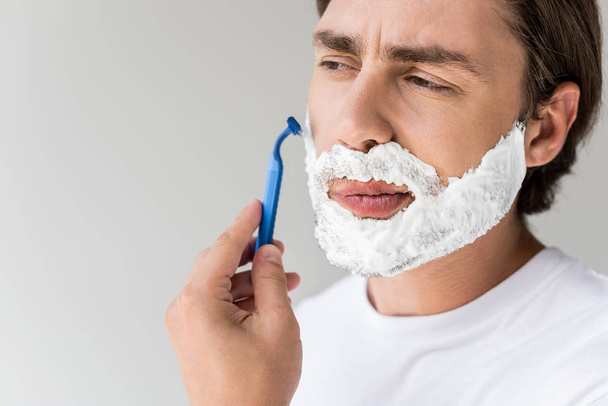 portrait of attractive man with foam on face and razor shaving beard isolated on grey - 写真・画像