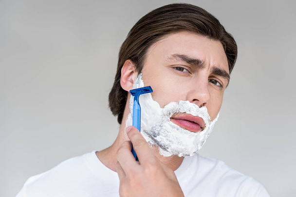 portrait of attractive man with foam on face and razor shaving beard on grey backdrop - Foto, immagini