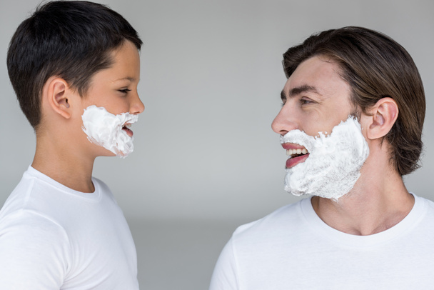 side view of happy father and son with shaving foam on faces on grey background - 写真・画像