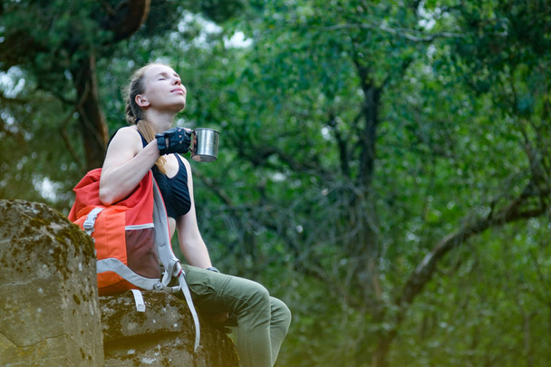 Mujer joven aventurera con mochila naranja de viaje sentado en una antigua roca fortaleza y beber café de la taza
.  - Foto, Imagen