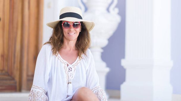 Μέση ηλικία μελαχρινή γυναίκα φοράει καπέλο το καλοκαίρι στις διακοπές με ένα χαρούμενο πρόσωπο στέκονται και να χαμογελούν με αυτοπεποίθηση χαμόγελο δείχνει τα δόντια - Φωτογραφία, εικόνα