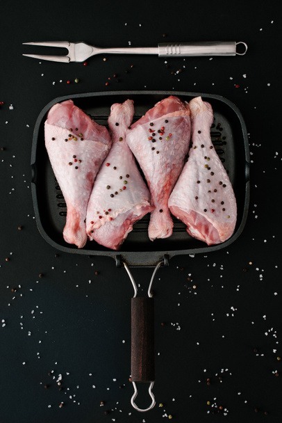 acostado plano con patas de pavo crudo con granos de pimienta y sal en la sartén con tenedor de carne en negro
 - Foto, Imagen