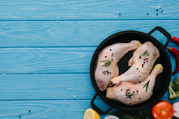 bovenaanzicht van de poten van de kip in de pan en verschillende groenten en specerijen op blauwe houten tafelblad - Foto, afbeelding