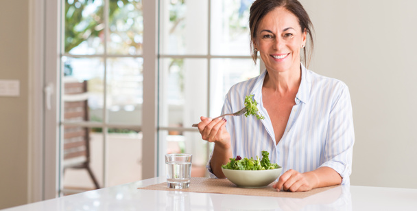 Middle aged woman eating fresh salad in a bowl at home with a happy face standing and smiling with a confident smile showing teeth - Foto, Imagen