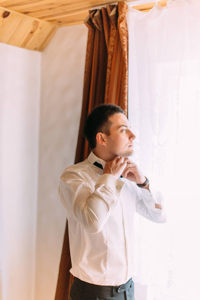 Half-length portrait of the groom tying the bow-tie while looking through the window. - Фото, зображення