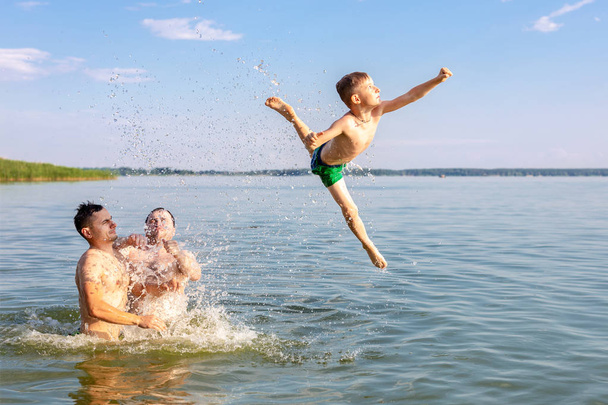 Twee jonge volwassenen en één kind jongen met plezier in de rivier- of meerwater. Kind hoog springen met hulp van vrienden. Zomer-concept voor outdoor activiteiten en recreatie - Foto, afbeelding