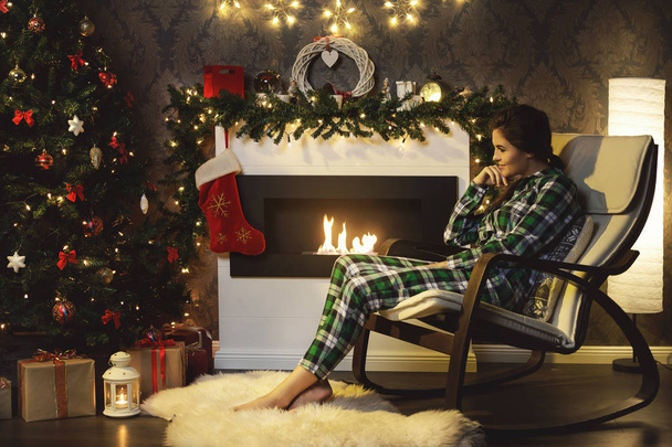 Christmas or New Year celebration. Happy woman sitting in the rocking chair beside fireplace. - Photo, Image