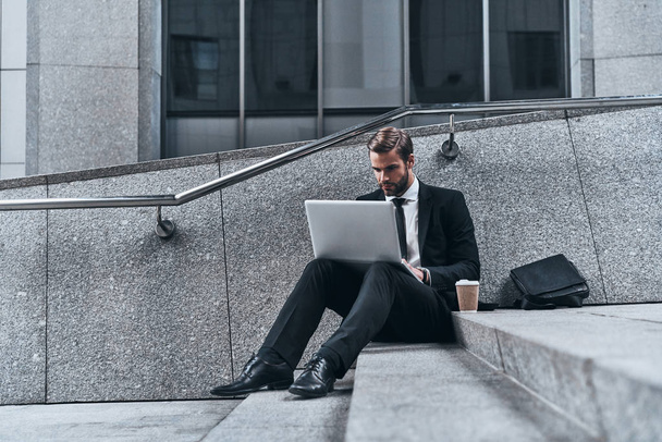 Using modern technologies, man in suit using laptop while sitting on stairs outdoors - Photo, image