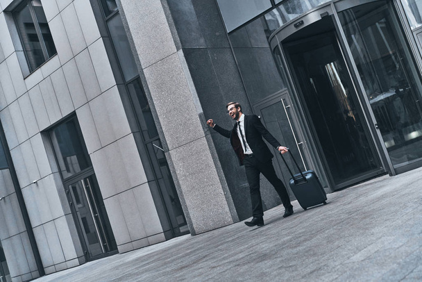 happy handsome businessman in suit pulling luggage while walking outdoors at office building - Valokuva, kuva