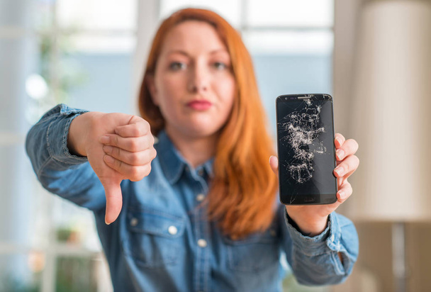 Mujer pelirroja sosteniendo teléfono inteligente roto con la cara enojada, signo negativo que muestra aversión con los pulgares hacia abajo, concepto de rechazo
 - Foto, imagen