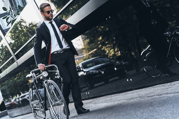 full length of handsome young man in full suit pulling his bicycle and checking time while walking outdoors - Fotoğraf, Görsel
