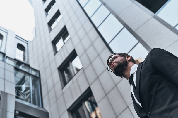 low angle view of good looking young man in full suit looking away while standing outdoors at office building - Fotoğraf, Görsel