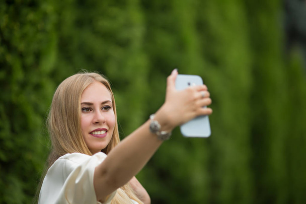 Young pretty blonde girl in a dress makes a selfie between thuja on the street in summer. - Foto, Imagen