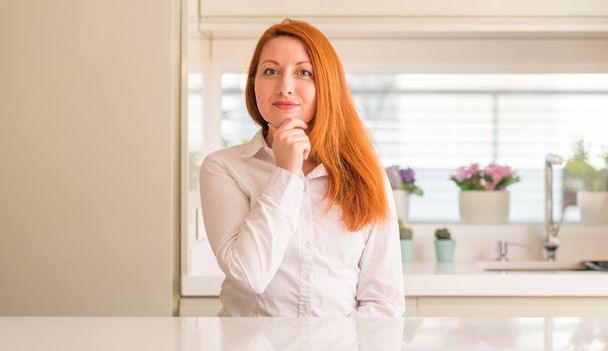 Redhead woman at kitchen looking confident at the camera with smile with crossed arms and hand raised on chin. Thinking positive. - Foto, afbeelding