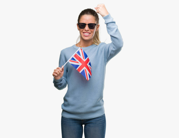 Mulher bonita segurando bandeira do Reino Unido irritado e frustrado gritando com raiva, louco e gritando com a mão levantada, conceito de raiva
 - Foto, Imagem