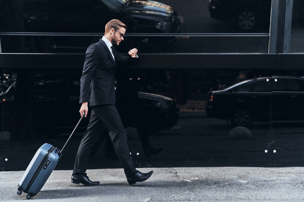 full length of young businessman in suit pulling luggage while walking outdoors - Valokuva, kuva
