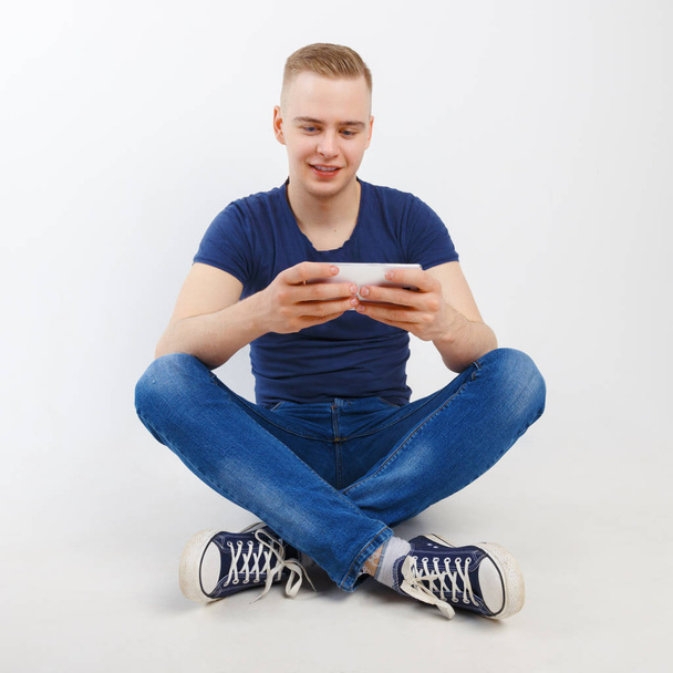 Young happy handsome man in blue shirt and jeans sits on floor with smartphone in white studio - Fotó, kép