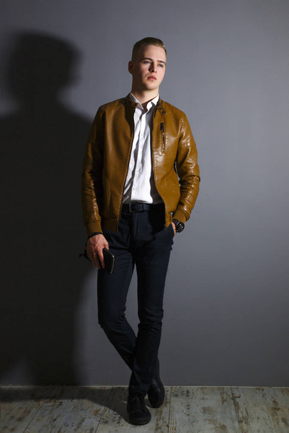 Όμορφος νεαρός άνδρας στο δερμάτινο jacket θέτει με ένα ΠΟΥΓΚΙ σε γκρι στούντιο, ολόκληρο το σώμα - Φωτογραφία, εικόνα