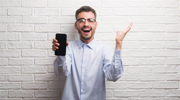 Young adult man talking on the phone standing over white brick wall very happy and excited, winner expression celebrating victory screaming with big smile and raised hands - Photo, Image